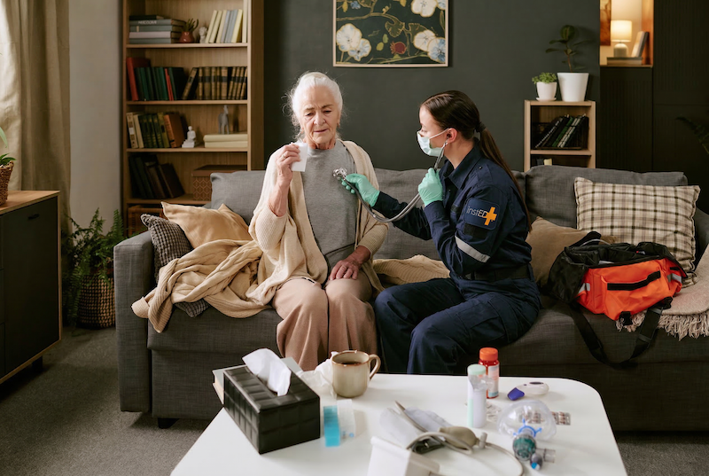 masked insted worker in an elderly womans home listening to her heart